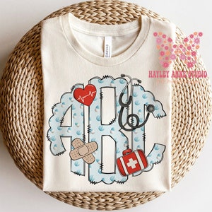 May include: A beige t-shirt with a blue and white monogram design featuring a stethoscope, a heart with a heartbeat line, a bandage, and a red first aid kit. The text "HAYLEY ANNE STUDIO" is visible in the top right corner of the image.