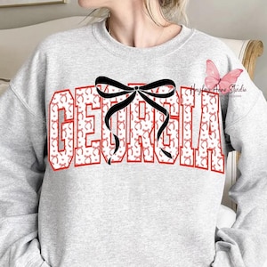 May include: A light gray sweatshirt featuring the word "GEORGIA" in red block letters with a pattern of red bows. A large black bow is centered over the word. The sweatshirt is a casual style.