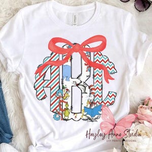 May include: White t-shirt featuring a colorful monogram design with the letters H, A, and K. The design includes a red bow, blue and red striped patterns, and illustrations of characters. The text "Hayley Anne Studio" is printed at the bottom.