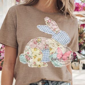May include: A tan t-shirt featuring a patchwork bunny design. The bunny is composed of floral, gingham, and lace fabrics in pastel colors. The design includes the text "Raspberry Anne Studio" and a butterfly graphic.