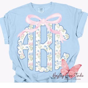 May include: Light blue t-shirt with a floral monogram and a pink bow. The monogram is surrounded by a scalloped edge. The text "Hailey Anne Studio" is in the bottom right corner.