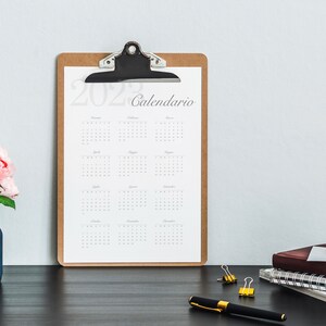 Può includere: Un portafoglio marrone con un calendario 2022 Calendario su una scrivania nera con un vaso di fiori rosa, una penna e graffette dorate.