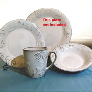 May include: A set of white ceramic dishes with a seashell design. The set includes a bowl, a plate, and a mug. The text "This plate not included" is written on the image.