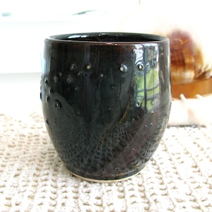 May include: A dark brown, cylindrical ceramic cup with a glossy finish and a series of small, scattered indentations across its surface. The base of the cup is unglazed, revealing a light beige clay color, while the rim is smooth and rounded.