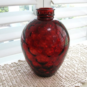 May include: A deep red glass vase with a rounded, bulbous body and a narrow, circular neck. The surface of the glass features a distinct, raised bubble or dimpled texture throughout, creating a tactile appearance with light reflecting off the curves.