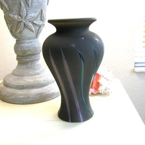 May include: A black ceramic vase with vertical purple and green painted accents. The vase has a rounded body and a flared rim. A decorative gray planter with starfish designs is in the background.
