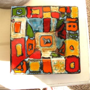 May include: Square ceramic dish with a colorful abstract design. The dish features a mosaic of shapes in red, orange, yellow, green, and blue, outlined in black. The dish is likely handmade and suitable for decorative use.