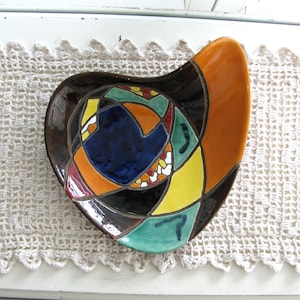 May include: A ceramic bowl with an irregular, asymmetrical shape featuring a glossy finish. The surface displays a segmented, stained-glass style pattern in vibrant shades of dark blue, bright yellow, orange, teal, and deep brown with black outlines.