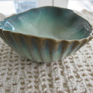 May include: A ceramic bowl shaped like a seashell with a scalloped edge. The exterior features a textured, ribbed surface with a gradient of earthy brown and deep teal tones, while the interior transitions to a smooth, pale turquoise glaze.