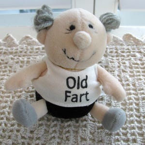 May include: A plush doll with a bald head, grey tufts of hair on the sides, and a round nose. The doll wears a white tank top with the text Old Fart printed in black, paired with black shorts and grey feet. The doll has a stitched smile.