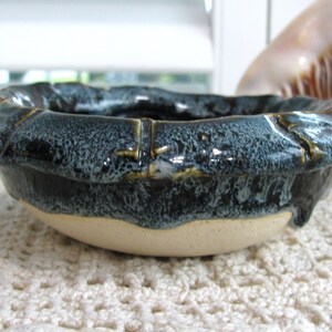 May include: A small, round ceramic bowl with a thick, textured rim featuring a dark, glossy blue and black glaze that drips down onto the unglazed, light beige base. The bowl has a segmented, bamboo-like appearance along the upper edge.