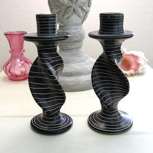 May include: Two black and white swirl design candle holders. The candle holders have a base, a twisted body, and a top. The candle holders are made of a dark material with white horizontal lines. A pink vase and a seashell are in the background.
