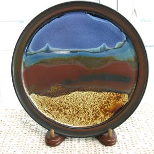 May include: A round ceramic plate featuring a multi-colored glaze with horizontal bands of deep blue, teal, reddish-brown, and a textured, speckled tan base. The plate has a dark brown rim and is displayed on a small wooden stand.