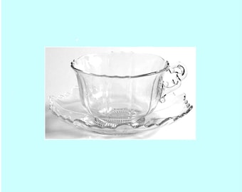May include: A clear glass teacup and saucer set featuring a scalloped edge design. The teacup has a curved handle and vertical fluted panels, while the matching saucer displays a coordinating scalloped rim and a textured base pattern.