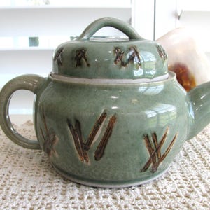 May include: A sage green ceramic teapot with a matching lid, featuring a rounded body, a curved handle, and a short spout. The surface is decorated with dark brown, incised, abstract linear markings across the body and the lid handle.