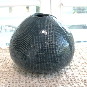 May include: A round, dark blue ceramic vase with a textured, woven fabric-like pattern across its entire surface. The vase has a glossy finish and a small, circular opening at the top with a slightly irregular, organic rim shape.