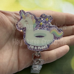 May include: A badge reel featuring a white unicorn floating on a pool float with a rainbow mane and tail. The badge reel has a glittery purple border and the text "I'm a float".