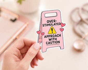 May include: A pink, A-frame floor sign sticker featuring a yellow caution triangle with an exclamation point. The text reads OVER-STIMULATED APPROACH WITH CAUTION. The design is decorated with small pink hearts on the sides of the sign.