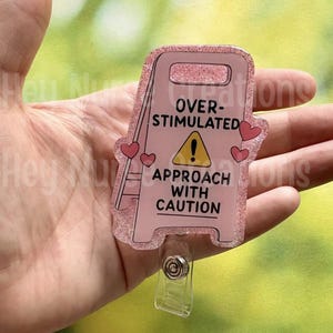 Könnte beinhalten: Ein rosafarbener, glitzernder Ausweishalter in Form eines Warnschildes. Der Text lautet OVER-STIMULATED APPROACH WITH CAUTION mit einem gelben Warndreieck-Symbol. Kleine rosa Herzen sind aufgedruckt und ein transparenter Clip ist unten befestigt.