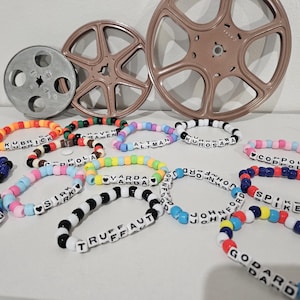 Pode incluir: Uma coleção de pulseiras de contas coloridas com os nomes de diretores de cinema escritos em contas de letras brancas. Bobinas de filme estão no fundo. As pulseiras são em várias cores, incluindo vermelho, azul, amarelo e preto.