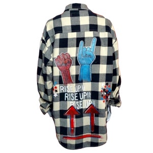 May include: A black and white plaid flannel shirt with red, blue, and white painted designs. The designs include two raised fists, a hand with a rock and roll hand gesture, and the words "Rise Up!" repeated three times. There are also red arrows pointing upwards.