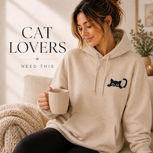 Può includere: Una felpa con cappuccio beige con una grafica di gatto nero sulla tasca. Il testo "CAT LOVERS NEED THIS" è visualizzato in nero. Una donna tiene in mano una tazza beige.