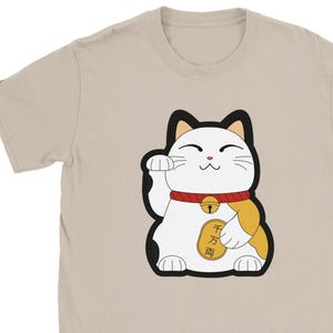 Könnte beinhalten: Beiges T-Shirt mit einem Cartoon-Design einer Maneki-neko-Katze. Die Katze ist weiß mit schwarzen Umrissen, einem roten Halsband und einer goldenen Glocke. Die Katze hebt ihre linke Pfote. Das Shirt ist aus weichem Material.