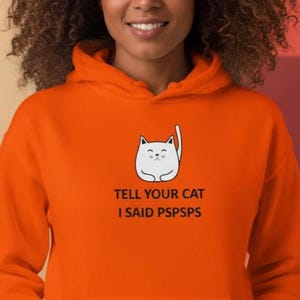 Könnte beinhalten: Leuchtend orangefarbener Kapuzenpullover mit einer weißen Cartoon-Katze und dem Text "TELL YOUR CAT I SAID PSPSPS". Der Hoodie hat eine Kapuze und eine Vordertasche.