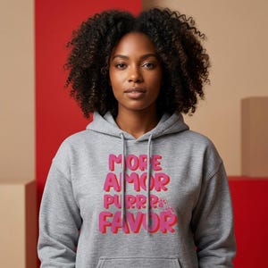 Könnte beinhalten: Ein grauer Kapuzenpullover mit den Worten "MORE AMOR PURRR FAVOR" in rosa und roten Blockbuchstaben. Der Pullover hat eine Kapuze mit Kordelzug und eine Vordertasche. Das Model trägt den Pullover.