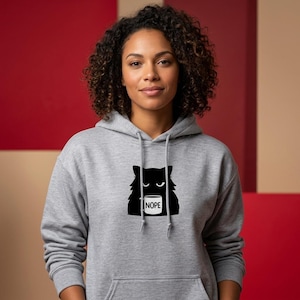 Könnte beinhalten: Grauer Kapuzenpullover mit einem schwarzen Katzenmotiv, das eine Tasse mit dem Wort "NOPE" hält. Der Pullover hat eine Vordertasche und eine Kapuze mit Kordelzug. Das Katzenmotiv ist mittig auf der Brust.