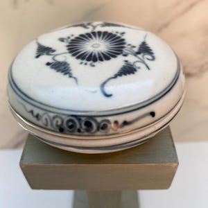 May include: A small, round, white ceramic box with a blue and black floral design. The box is sitting on a wooden stand.