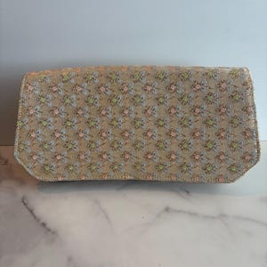 May include: A rectangular clutch with a beige background and a repeating floral pattern in pink, green, and blue. The clutch has a slightly rounded top and a light-colored border.