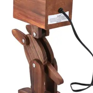 May include: A wooden robot lamp with a brown finish. The lamp has a rectangular wooden base and a jointed arm that can be adjusted. The lamp is plugged in and has a black cord.