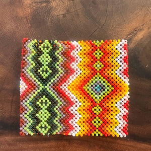 Può includere: Una toppa quadrata di perline con un motivo geometrico a rombi e zig-zag. Il design incorpora colori vivaci, tra cui perline di vetro verde lime, verde bosco scuro, giallo brillante, arancione, rosso e bianco disposte in una griglia.