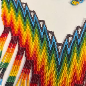 May include: A beaded necklace featuring a vibrant chevron pattern in rainbow colors including red, orange, yellow, green, blue, and dark brown. The design incorporates small white accents within the zig-zag rows and includes long, dangling beaded fringe strands.