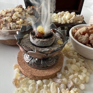 A display of burning copal resin on a charcoal disc in a decorative metal incense holder, producing thick aromatic smoke.