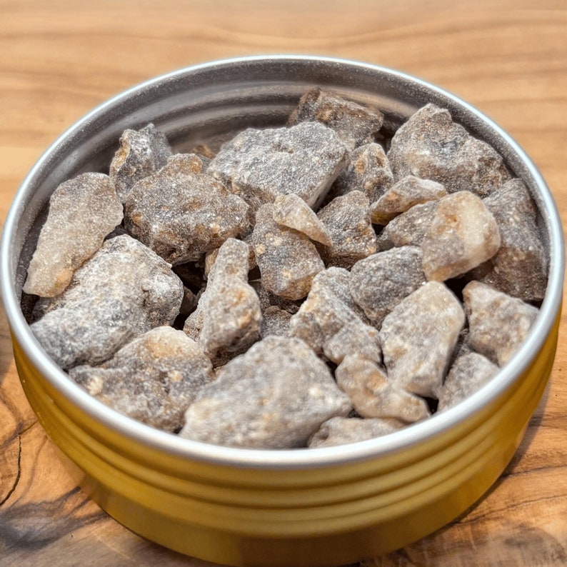 Sumatra Benzoin resin tears, showcasing rough, grayish-brown pieces with a natural, rugged texture. The resin chunks have a slightly translucent quality, emphasizing their raw, earthy appearance on the wooden surface beneath.