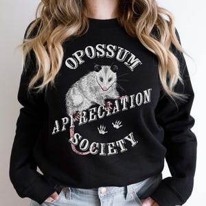 A woman wearing a black sweatshirt. The sweatshirt features an opossum and the words Opossum Appreciation Society. The opossum is sitting on the word Appreciation.