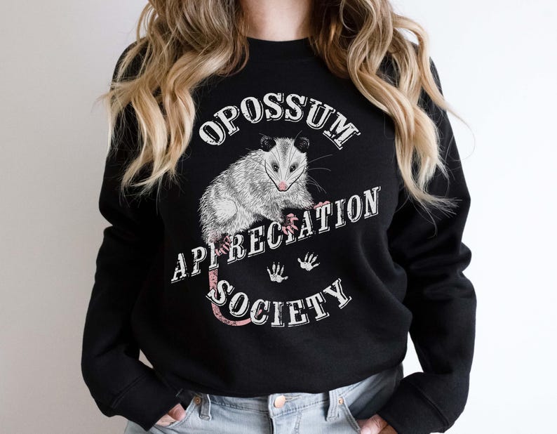 A woman wearing a black sweatshirt. The sweatshirt features an opossum and the words Opossum Appreciation Society. The opossum is sitting on the word Appreciation.