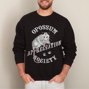 A man wearing a black sweatshirt. The sweatshirt features an opossum and the words Opossum Appreciation Society. The opossum is sitting on the word Appreciation.