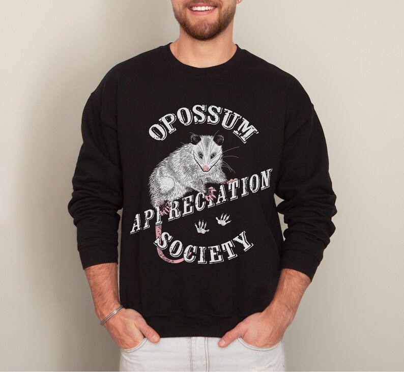 A man wearing a black sweatshirt. The sweatshirt features an opossum and the words Opossum Appreciation Society. The opossum is sitting on the word Appreciation.