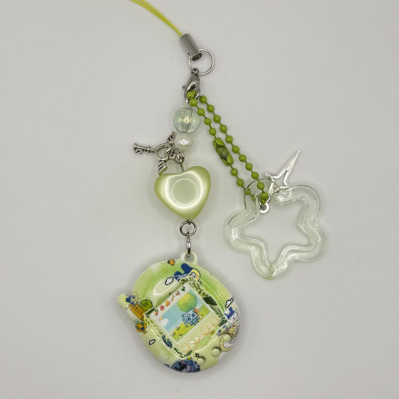 Pu&ograve; includere: Un portachiavi per animali virtuali verde lime con un guscio stampato con grafiche blu e gialle. Il ciondolo include una perla a forma di cuore verde chiaro, una piccola chiave argentata, un pendente a stella trasparente e una catenella a sfere verde.