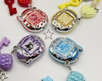 Könnte beinhalten: Vier Tamagotchi-Schlüsselanhänger mit den Charakteren Hello Kitty, Pompompurin, Kuromi und Cinnamoroll auf den Bildschirmen. Jedes Gerät ist mit einem farbigen Schlüsselanhänger, einer herzförmigen Perle und einem kleinen Stern an einer Kette versehen.