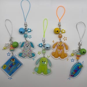 May include: Five phone charms featuring acrylic Webkinz characters and logos. Each charm includes a colorful cord, a small bell, star-shaped beads, and round beads. The charms display various dog illustrations and the Webkinz logo with the text discover a virtual world.