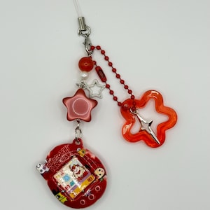 May include: A red Tamagotchi Paradise digital pet device attached to a beaded charm featuring a red star bead, a small white pearl, a silver star charm, a red ball chain, and a translucent orange flower-shaped charm with a silver star accent.