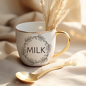 May include: White ceramic mug with a gold rim and a gold leaf design. The mug says "MILK" in black lettering.