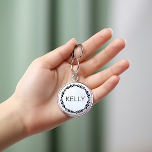 May include: A silver keychain with a round tag that says "KELLY" in black lettering surrounded by a black wreath design.