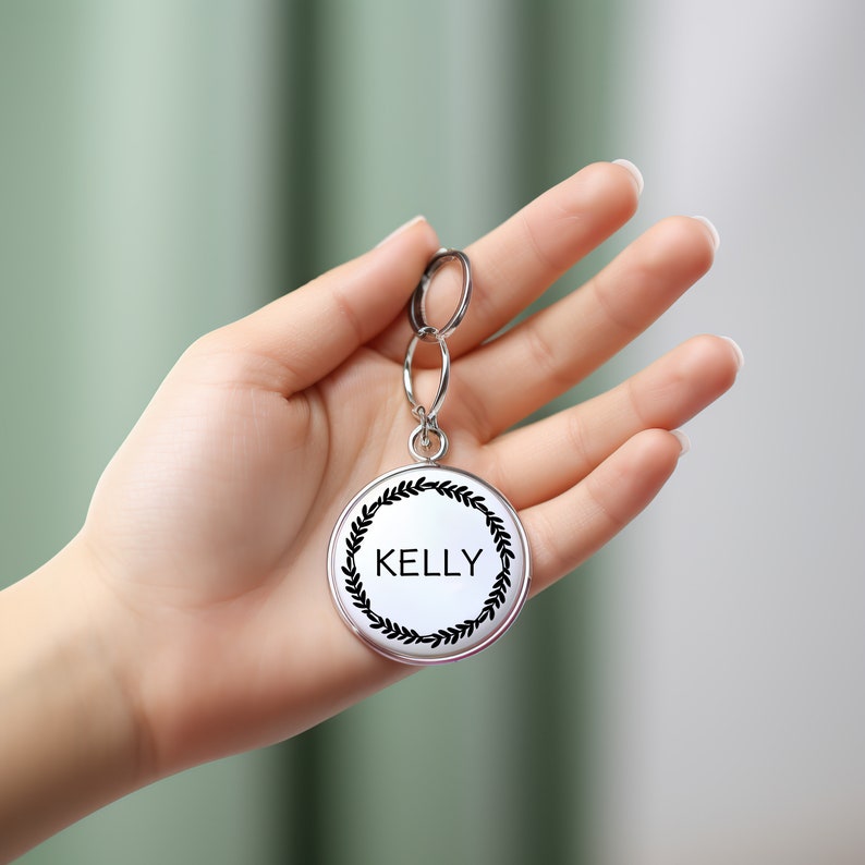 May include: A silver keychain with a round tag that says "KELLY" in black lettering surrounded by a black wreath design.
