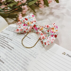 May include: A floral fabric bow with a gold-colored metal bookmark is placed on an open book. The bow features a white background with small red, pink, yellow, and blue flowers. Pink flowers are in the background.