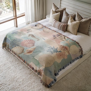 May include: A woven throw blanket featuring a watercolor design of seashells, seahorses, and coral in pastel colors. The blanket has a fringed edge and is displayed on a bed with pillows.